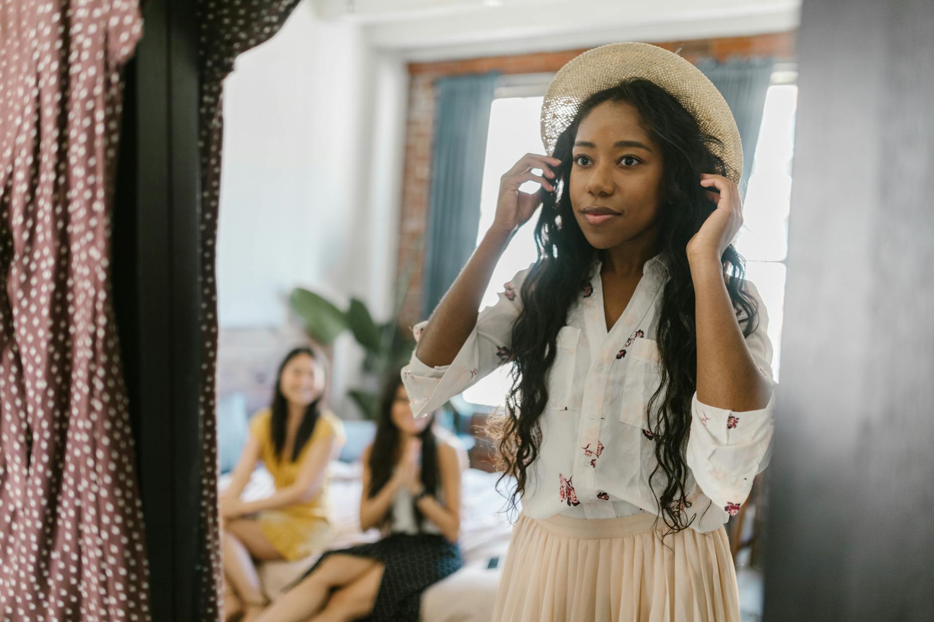 Młoda kobieta przymierza modny kapelusz przed lustrem z koleżankami w tle